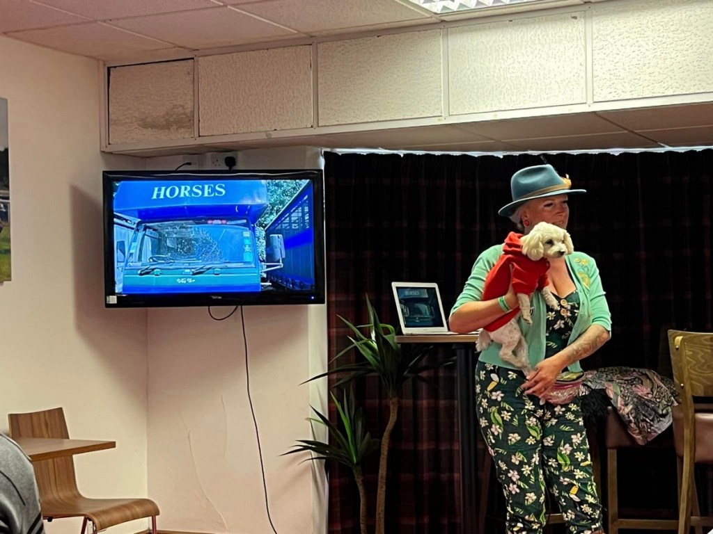 Nicole and her small white pup Sandy giving a talk at the Overland Show with a slide showing the blue horsebox they converted into their home