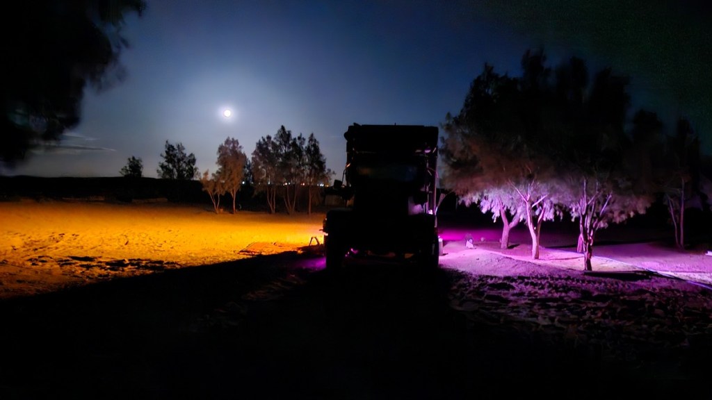 Image of Jacqueline Lambert's truck, The Beast, in the Sahara Desert with a full moon rising behind and coloured lights shining beneath the truck.
